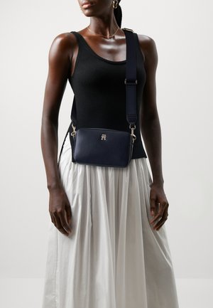 Borsa a tracolla in pelle blu navy con hardware dorato, forma rettangolare, superficie texturizzata, tracolla regolabile, presenta in modo prominente il logo sul fronte.
