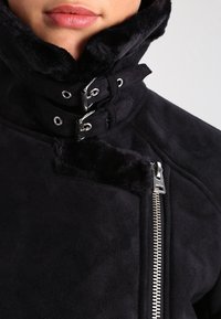 Svart fuskpälsjacka med hög krage, med silverdetaljer, en ficka med dragkedja och mjuk textur längs kragen och fram.