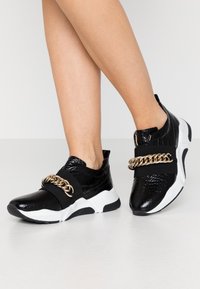 Baskets synthétiques noires avec une texture crocodile glossy, présentant un accent en chaîne dorée et une semelle blanche. Bandes noires extensibles pour un ajustement sécurisé.