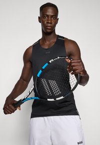 Svart treningssinglett med grå detaljer, iført hvite shorts. Subjektet holder et tennisslag med et svart og blått håndtak.