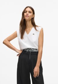 Λευκό αμάνικο τοπ με V λαιμόκοψη και μικρή γραφική λεπτομέρεια στο στήθος, σε συνδυασμό με μαύρους παντελόνες ψηλής μέσης με γκρι ζώνη.