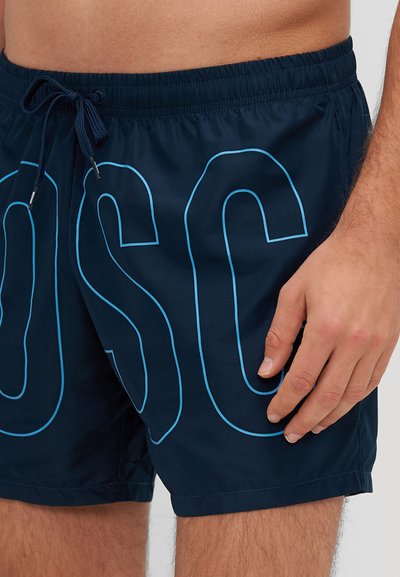 Jūras spēku peldšorti ar gaiši zilu pārmērīgi lielu "OSC" izdrukā, elastīgu vidukļa joslu ar auklu un gludu, ātri žūstošu audumu.