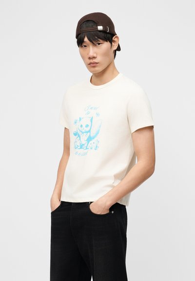 Jeune homme portant un t-shirt crème avec un motif de chat bleu, un pantalon noir et une casquette marron portée à l'envers, debout les mains dans les poches.