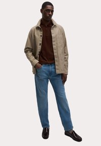 Chaqueta de algodón beige con cierre de botones y bolsillo, llevada sobre una camisa de manga larga marrón, combinada con jeans de color azul claro y mocasines oscuros.