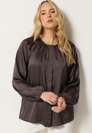 Blonde Frau trägt eine dunkle Satinbluse mit langen Ärmeln, gerafftem Ausschnitt und Bündchen, steht vor einem einfarbigen hellen Hintergrund.