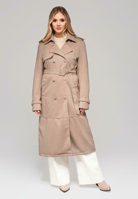 Beżowy dwurzędowy trencz z paskiem, wykonany z teksturowanej tkaniny, z pagonami na ramionach i szerokimi rękawami, zestawiony z białymi spodniami.