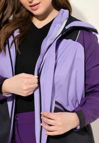 Lila och svart zip-up jacka med huva, med en blandning av matta och glänsande texturer, och en fleece-fodrad insida runt kragen.