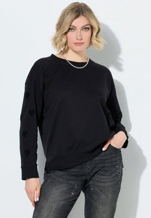 Ung kvinde med kort blondt hår iført en sort langærmet top med hjerte-detaljer og mørke jeans med rhinsten-accenter.