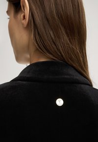 Donna con capelli castani che indossa una giacca di velluto nero, mostrando un piccolo distintivo metallico rotondo con un logo di cervo e il testo "JOOP!" sul retro.