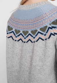 Gros plan sur une personne portant un pull en tricot gris avec des motifs en zigzag et en cœur pastel bleu, rose et marine sur le yoke.