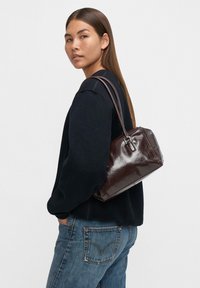 Kobieta z długimi brązowymi włosami, ubrana w czarny sweter, niebieskie dżinsy i nosząca ciemnobrązową skórzaną torebkę na ramieniu.