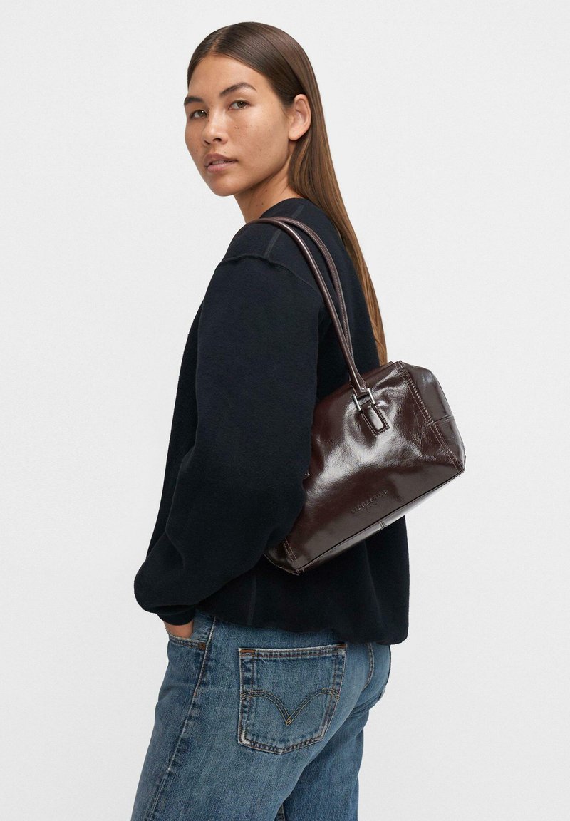 Kobieta z długimi brązowymi włosami, ubrana w czarny sweter, niebieskie dżinsy i nosząca ciemnobrązową skórzaną torebkę na ramieniu.