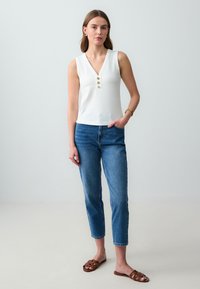 Witte geribbelde tanktop met een v-hals en drie decoratieve gouden knopen, gecombineerd met lichtblauwe slimfit jeans en bruine slidesandalen.
