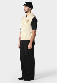 Beige pletená bezrukávová svetr se vzorem copánků a květinovou výšivkou, nošený přes černou košili s krátkým rukávem, kombinovaný s černými kalhotami a čepicí.