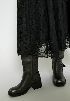 Černé kožené vysoké boty s postranním zipem, detaily pásku a širokým podpatkem, spárované s krajkovou sukní, která ukazuje složité vzory.