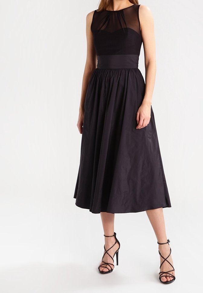 Abito nero caratterizzato da un corpetto aderente con sovrapposizione trasparente, una larga vita e una gonna ampia. Abbinato a sandali neri con cinturini.