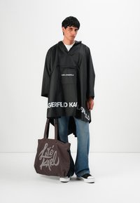 Man in zwarte Karl Lagerfeld regenponcho, blauwe jeans, zwart-witte sneakers, met een bruin totebag met de tekst "Love Karl".