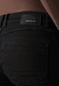 Zwarte jeans met een gladde textuur, voorzien van een zwartleren label met de opschrift "MOS MOSH" en stikseldetails op de zakken.