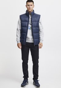 Meripadi takki tummansinisena harmaata hupparia, mustia housuja ja sinisiä lenkkareita. Takissa on korkea kaulus ja pieni logo rinnassa.