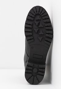 Zwarte rubberen schoenzool met een gestructureerd oppervlak en een diep profielpatroon. Voorzien van driehoekige noppen voor grip en een afgerond hielgebied.