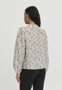 Bluse mit floralem Muster, langen Ärmeln, hellem Creme-Hintergrund und dunklen Akzenten. Sie hat einen runden Ausschnitt und geraffte Schultern.