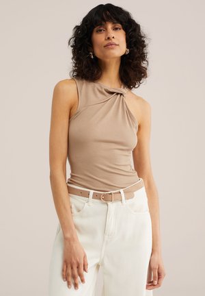 Donna con capelli ricci e scuri che indossa un top beige senza maniche con dettaglio a nodo e pantaloni bianchi a vita alta con cintura beige, in piedi davanti a uno sfondo neutro.