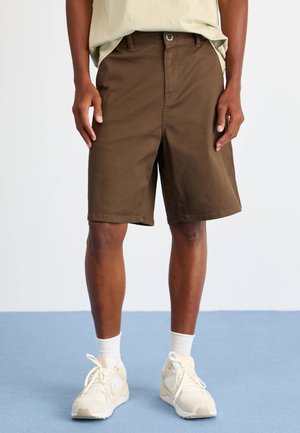 Shorts marron à coupe droite, avec une fermeture à bouton et des poches latérales. Portés avec des chaussettes blanches et des baskets claires sur un sol bleu.