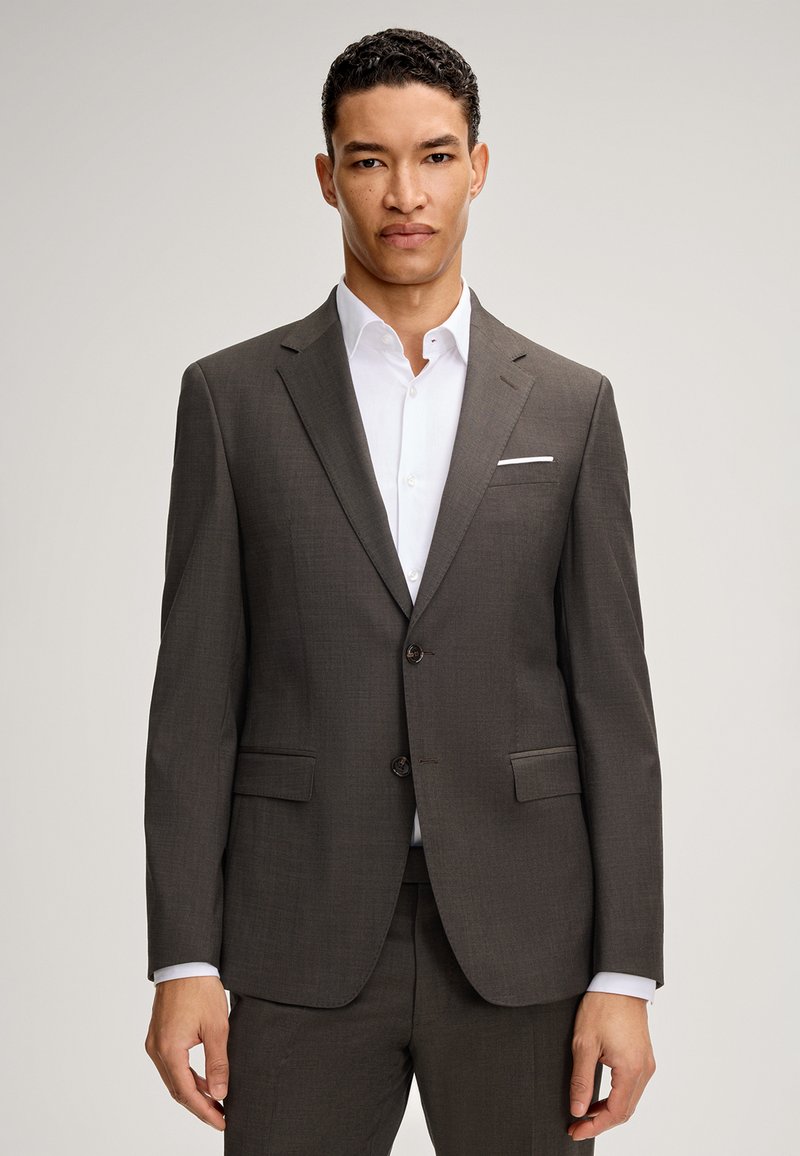 Completo grigio scuro con un tessuto strutturato. Presenta un risvolto a punta, due tasche frontali, una chiusura a singolo bottone e una camicia bianca sotto.