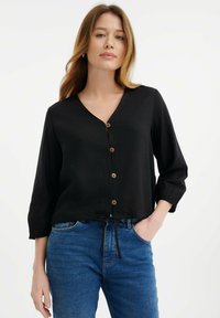 Zwarte overhemd met V-hals en lange mouwen, voorzien van houten knopen, een strik in de taille en een relaxte pasvorm. Denim jeans zijn zichtbaar.