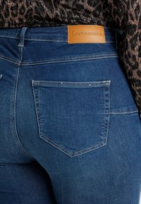 Primer plano del bolsillo trasero de unos jeans de mezclilla azules con una etiqueta de cuero marrón de la marca Junarose y parte de una blusa marrón con patrón.