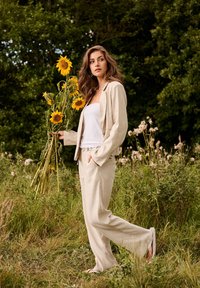 Femme en costume beige tenant de grandes tournesols marchant à travers un champ herbeux avec des arbres en arrière-plan.