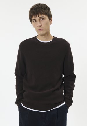 Brauner Strickpullover mit gerippter Textur, Rundhalsausschnitt und langen Ärmeln, über einem weißen Hemd getragen. Dunkelblaue Cordhosen.