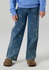 Šviesiai mėlynas megztinis derinamas su plačiomis džinsinių kelnių detalėmis, puoštomis sidabrinėmis širdies formos papuošalais, vidutinio tono su klasikine džinsų tekstūra.