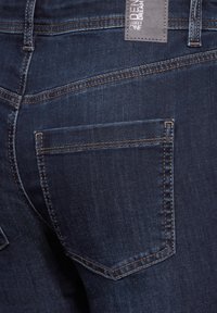 Jeans en denim bleu foncé avec une poche arrière ornée de surpiqûres oranges et une étiquette sur la ceinture. Le tissu semble lisse avec une légère texture.