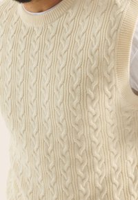 Krämfärgad kabelstickad ärmlös tröja med rund halsringning och strukturerad mönstring, tillverkad av mjukt stickad tyg.