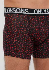 Boxer briefs neri con un motivo astratto rosso. Il materiale sembra morbido con una texture liscia. Il cinturino è nero con un marchio bianco.