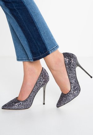 Glitrende sølvfarvede høje hæle sko med spids tå og sølvfarvet stiletthæl, kombineret med blå denim cropped bukser med farveblokering.