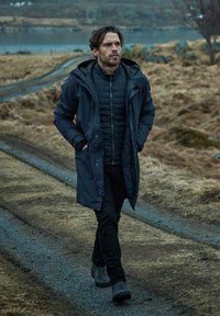 Man som går på en grusig stig, iförd en mörkblå vattentät parka, en svart puffjacka under, svarta byxor och svarta kängor.