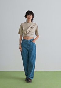 Jeune personne avec des cheveux bouclés courts portant un t-shirt court noué beige, un jean taille haute bleu et des baskets foncées, se tenant sur un sol vert.