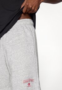 Γκρι βαμβακερές φόρμες με ελαστική μέση, πλαϊνές τσέπες και κόκκινο κεντημέν ο λογότυπο "Champion" στο αριστερό πόδι.