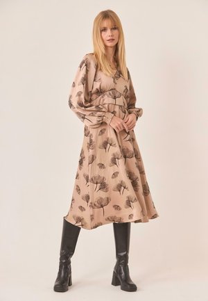 Mujer con el cabello rubio, vistiendo un vestido midi beige con flores, mangas largas abullonadas y botas negras de tacón hasta la rodilla, de pie frente a un fondo liso.