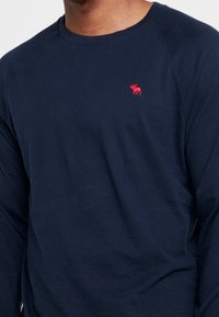 Marineblaue Langarm-T-Shirt aus Baumwolle mit Raglanärmeln und einem kleinen roten Elch-Logo auf der linken Brust. Weicher Stoff, lässige Passform.