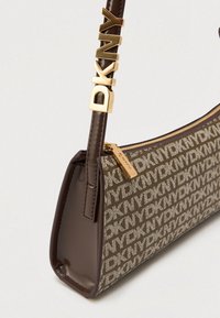 Καφέ τσάντα χειρός DKNY με μονογράμματα, δερμάτινες πλευρές, χρυσό φερμουάρ και χρυσά γράμματα DKNY στα κρεμαστάρια.