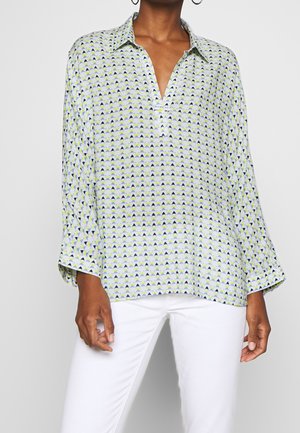 Femme portant un chemisier ample à manches longues à motif avec un col et un pantalon blanc, debout devant un fond uni.