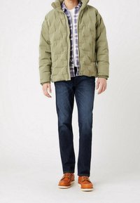 Chaqueta acolchada de color verde oliva con cierre de cremallera, con capucha, llevada sobre una camisa de cuadros, combinada con jeans de mezclilla oscura y zapatos marrones.