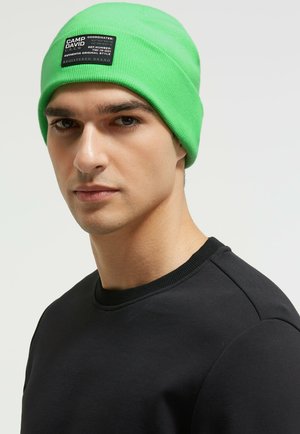 Green knit beanie with a black rectangular label featuring white text. Worn with a black crewneck shirt. Smooth texture, closely fitting design.