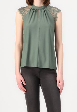 Woman wearing sleeveless green top with lace shoulder details and keyhole neckline, paired with dark fitted pants.