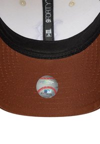 Gorra de béisbol marrón con visera negra y una textura de tela suave. Un adhesivo circular de la MLB con un detalle de costura roja está fijado a la parte inferior.