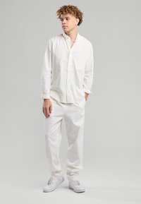 Giovane con capelli ricci che indossa una camicia bianca larga, pantaloni bianchi e sneakers bianche, in piedi con una mano in tasca guardando di lato.