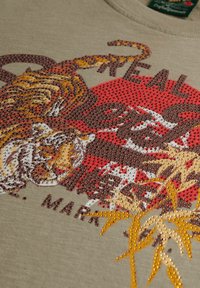 Camiseta beige con un gráfico de tigre colorido adornado con strass, acentuado por detalles rojos y amarillos, y una textura de tela suave.